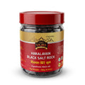 A jar of Vajra Foods Himalayan Black Salt Rock, featuring a red lid and a red and gold label, set against a plain white background.